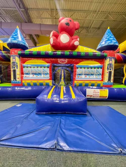 A colorful inflatable bounce house with a large teddy bear on top and a slide in front.