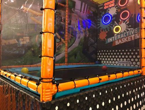 A colorful interactive basketball game inside a trampoline park, featuring a netted enclosure and bright lights.