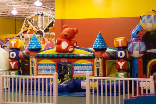 A colorful inflatable bounce house with a teddy bear on top, surrounded by a white fence in a play area.