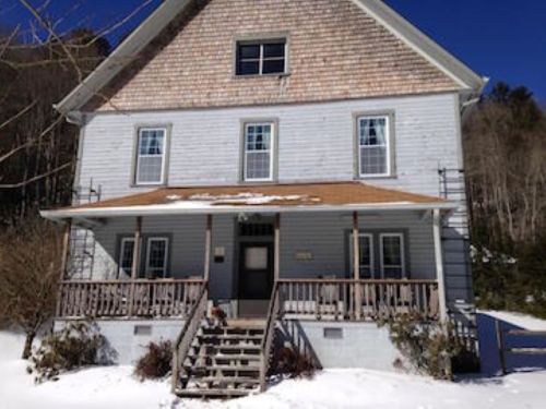 A three-story house with a wooden porch, surrounded by snow and trees, under a clear blue sky.
