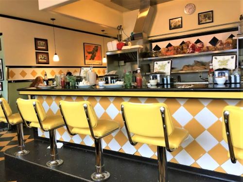 A retro diner interior featuring yellow bar stools, a counter with condiments, and vintage decor on the walls.