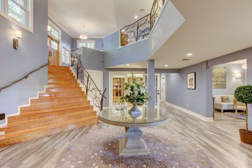 A spacious foyer with a wooden staircase, round table with flowers, and stylish decor in soft blue tones.