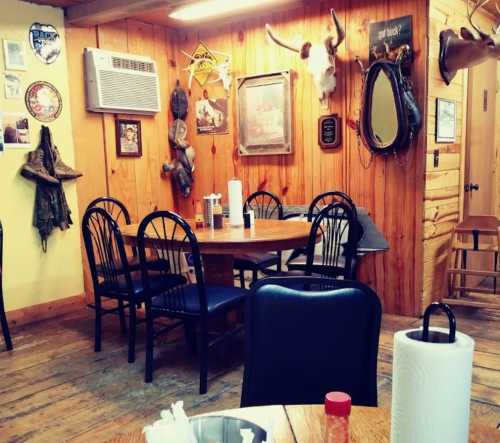 Cozy restaurant interior with wooden walls, a round table, chairs, and rustic decor including animal mounts and signs.