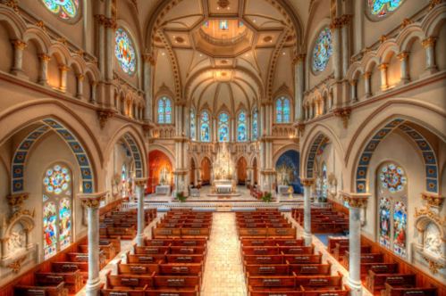 Interior of a grand church featuring stained glass windows, ornate arches, and wooden pews arranged in rows.