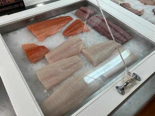 A display of various fish fillets on ice, covered by a glass top, showcasing salmon and white fish varieties.