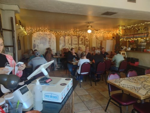A cozy restaurant interior with patrons dining, string lights overhead, and a counter with a register in the foreground.