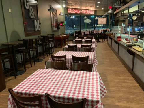 A cozy restaurant interior with checkered tablecloths, wooden chairs, and a counter displaying food options.