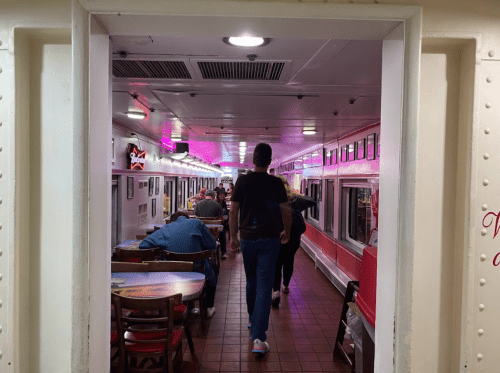 A view of a brightly lit restaurant hallway with diners at tables and a person walking towards the camera.