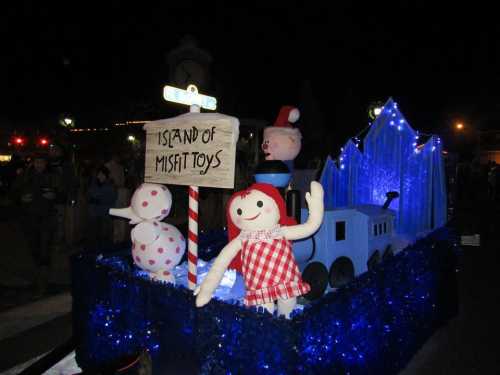 A festive float featuring oversized toys, including a doll in a red dress, snowmen, and a train, with a sign reading "Island of Misfit Toys."