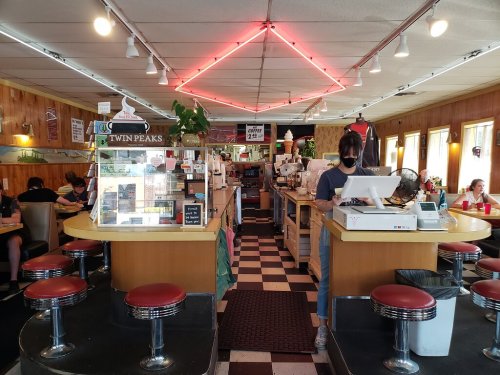 A retro diner interior with checkered floors, red stools, and a counter where a masked employee takes orders.