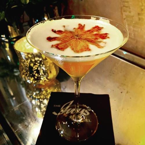 A cocktail in a martini glass topped with a dried flower, set on a reflective surface next to a lit candle.