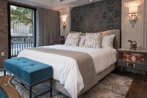 A stylish hotel room featuring a plush bed, decorative pillows, a blue ottoman, and elegant wall decor.