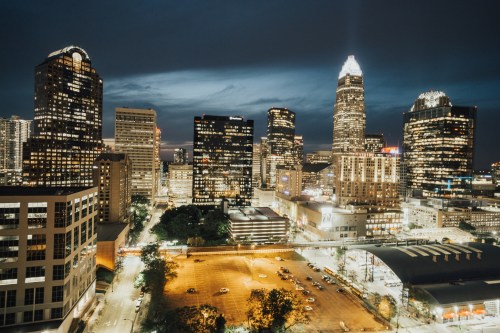 City skyline at night with illuminated buildings and a parking lot below, showcasing a vibrant urban atmosphere.