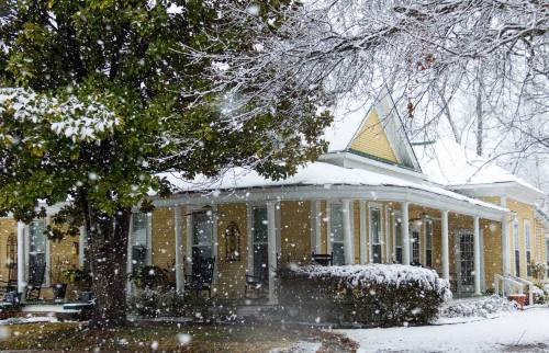 A charming yellow house with a snow-covered roof, surrounded by falling snowflakes and a large tree.