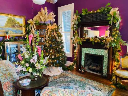 Cozy living room with a purple wall, decorated Christmas tree, floral arrangements, and a fireplace adorned with greenery.
