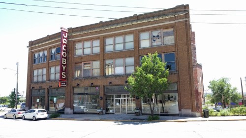 Historic brick building with large windows and a sign reading "Jacoby's," surrounded by trees and parked cars.