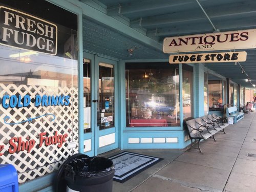 A storefront featuring signs for "Fresh Fudge," "Antiques," and "Cold Drinks," with a bench and trash can outside.