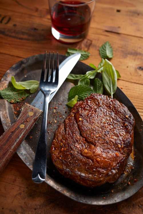 A juicy steak on a metal plate, garnished with fresh mint leaves, alongside a fork and knife, with a glass in the background.