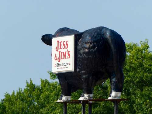 A large black cow statue with a sign reading "Jess & Jim's Steakhouse" on its side, set against a blue sky.
