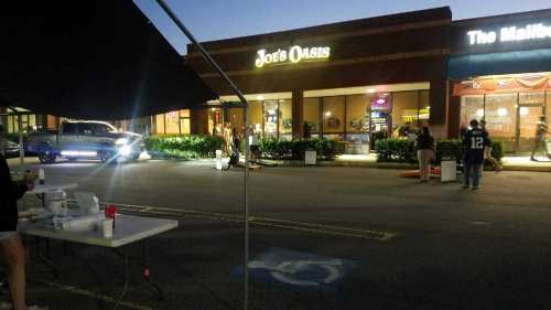 A street view at dusk featuring Joe's Oasis and The Mailbox store, with people and a table in the foreground.