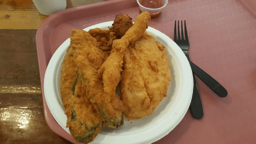 A plate of fried fish, chicken, and a side of curly fries, served with a small cup of dipping sauce.