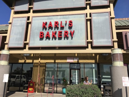 Exterior of Karl's Bakery with a bright sign, featuring a woman standing outside and plants in the foreground.