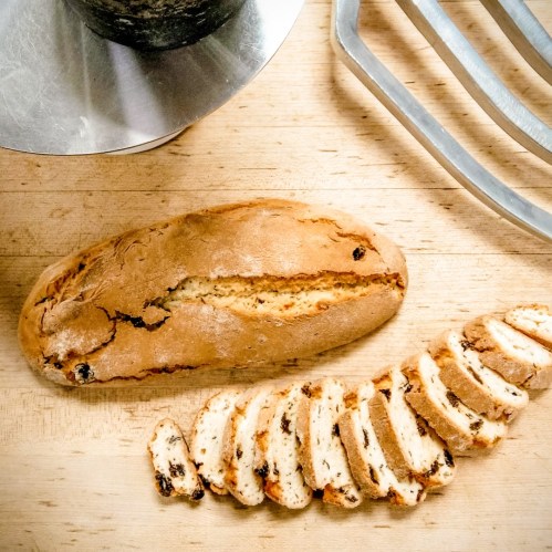 A loaf of bread with a few slices cut, resting on a wooden surface next to a metal mixing bowl and a whisk.