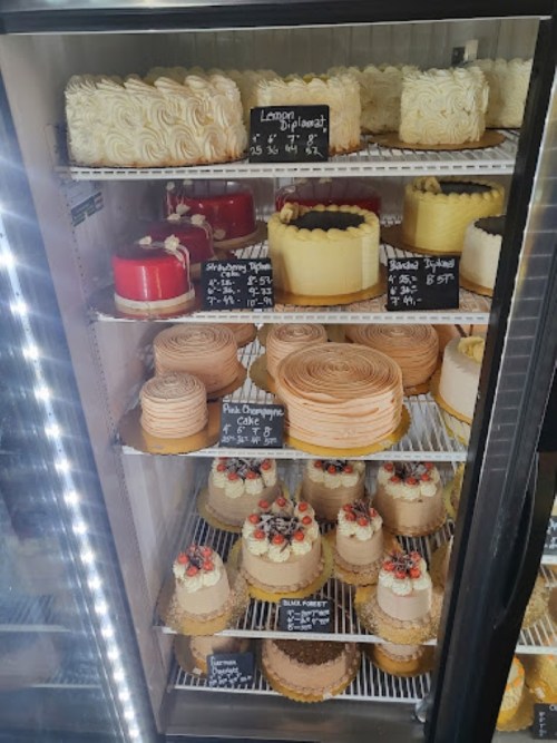 A display case filled with various cakes, including layered, frosted, and decorated options, each labeled with prices.