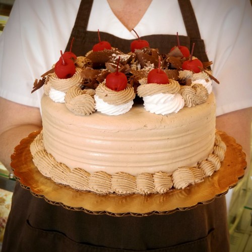 A person in an apron holds a chocolate cake topped with whipped cream, cherries, and chocolate pieces on a gold platter.