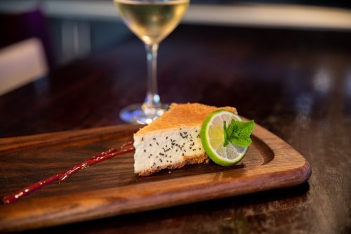 A slice of cheesecake with a lime wedge and mint on a wooden plate, accompanied by a glass of white wine.