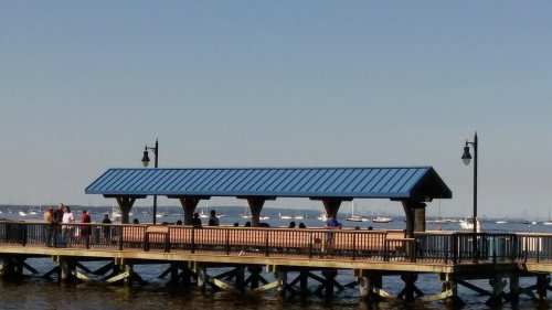 A wooden pier with a blue roof, people enjoying the view of the water and boats under a clear sky.