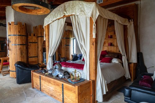 A rustic bedroom featuring a wooden canopy bed, armor, and medieval decor, with stone flooring and warm lighting.