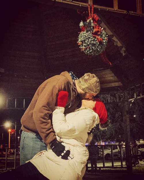 A couple shares a kiss under mistletoe, surrounded by festive decorations in a cozy winter setting.