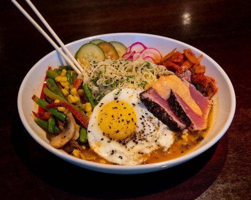 A bowl of colorful stir-fried vegetables, topped with a fried egg, seared tuna, and drizzled with sauce.