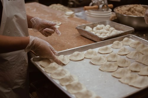 A person in gloves arranges uncooked dumplings on a baking tray in a kitchen setting.