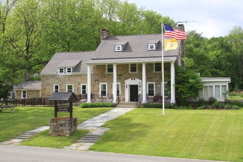 A large stone house with a front porch, American flag, and well-maintained lawn surrounded by trees.