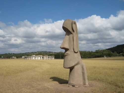 A large stone statue resembling a Moai stands in a grassy field under a cloudy sky, with distant stone structures visible.