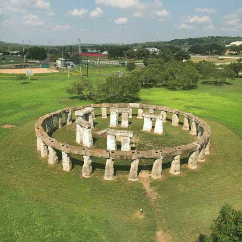 A circular stone structure resembling Stonehenge, surrounded by green grass and trees under a blue sky.