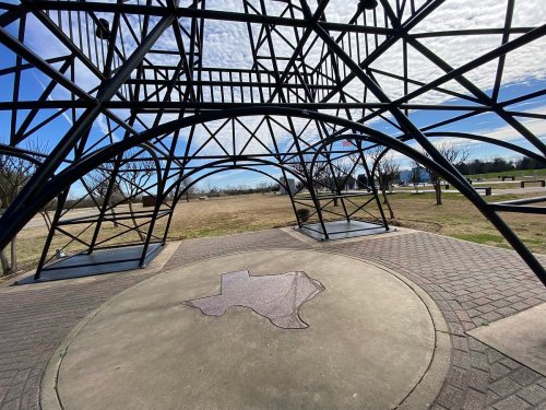 A metal structure with an open design, featuring a Texas outline on the ground, set in a grassy park under a blue sky.