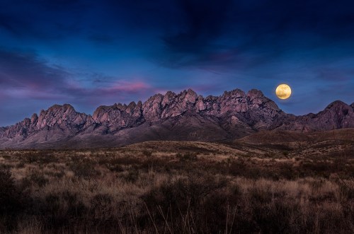 A dramatic mountain range under a twilight sky, with a large full moon rising above the peaks.
