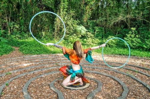 A person in colorful attire performs with hula hoops in a forested labyrinth, surrounded by lush greenery.