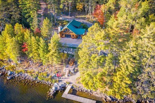 Aerial view of a cabin by a lake, surrounded by colorful trees and a wooden dock along the shoreline.