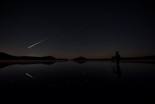 A silhouette of a person photographing a meteor streaking across a starry night sky over a calm lake.