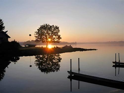 A serene lake at sunrise, featuring a silhouetted tree and calm water reflecting the colorful sky.