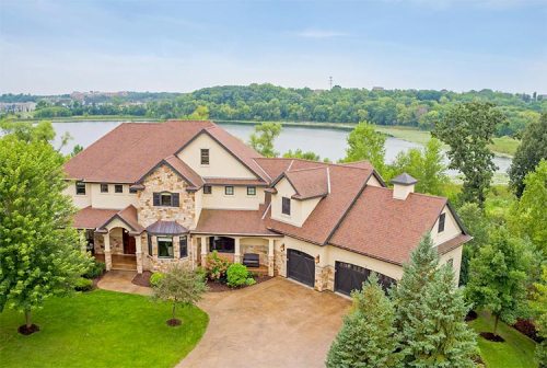 A beautiful two-story house with stone accents, surrounded by greenery and overlooking a serene lake.
