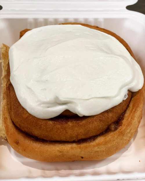 A freshly baked cinnamon roll topped with a thick layer of cream cheese frosting in a takeout container.