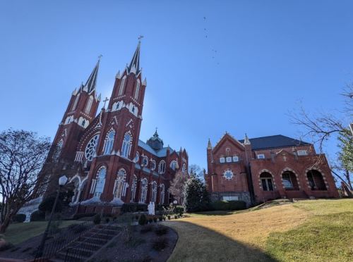 A large red brick church with tall spires, surrounded by greenery and a clear blue sky.