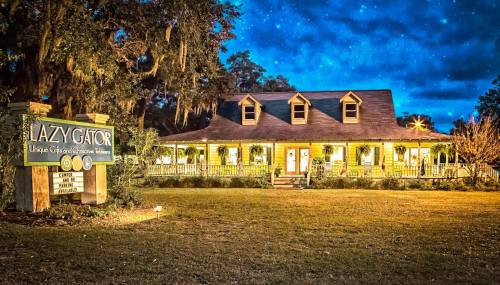A charming yellow house with a porch, illuminated at night, surrounded by trees and a sign reading "The Lazy Gator."
