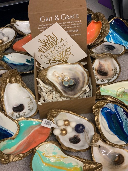 A collection of colorful, handcrafted oyster shell dishes with a gift box labeled "Grit & Grace" in the center.