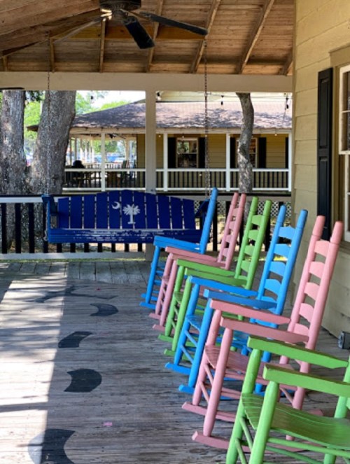Colorful rocking chairs in pink, blue, and green lined up on a porch with a ceiling fan and wooden beams above.